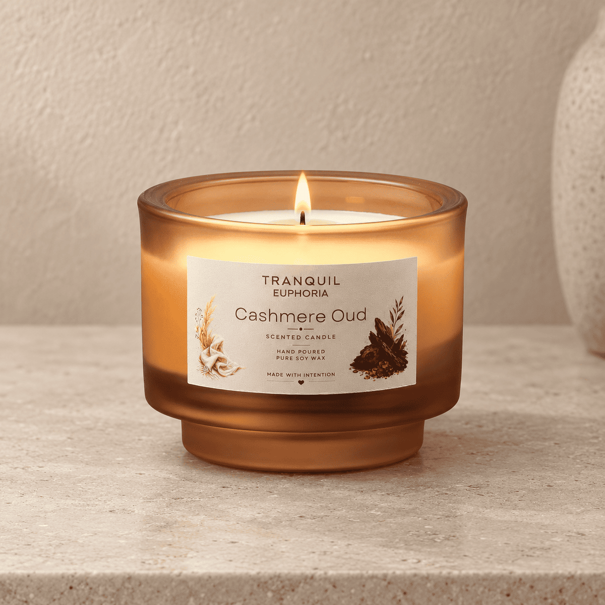 A lit Tranquil Euphoria Cashmere Oud scented candle in a translucent amber glass jar, set on a textured beige stone surface against a neutral beige background.