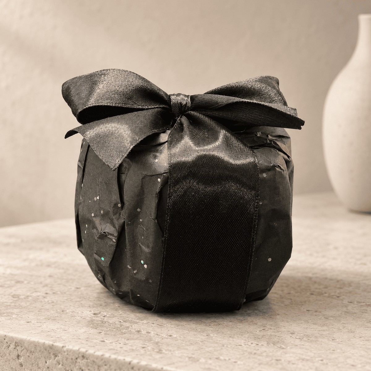 A small Midnight Sky candle  wrapped in black glittery paper with a large black satin bow, sitting on a light beige stone surface with a neutral background and a white vase partially visible.