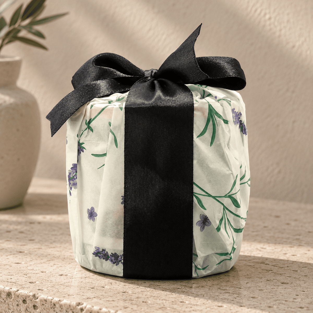A Tranquil Euphoria Oakmoss Aura candle wrapped in off-white tissue paper with a green floral pattern and purple flowers, tied with a wide black satin ribbon and bow, set on a stone surface.