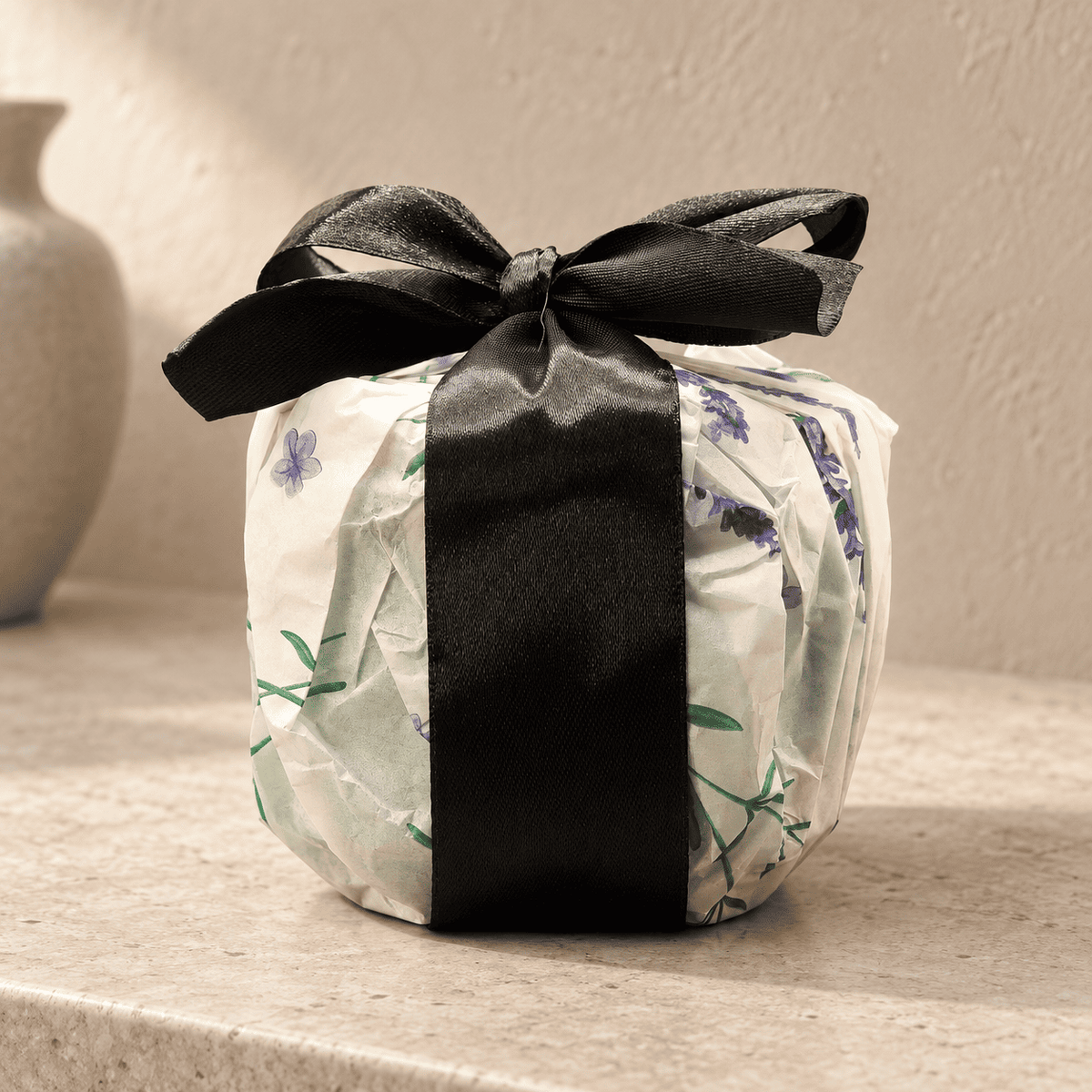 A gift-wrapped Oakmoss Aura package tied with a wide black satin ribbon bow, set on a beige stone surface against a textured beige wall. The wrapping paper is off-white with a delicate green botanical print and small purple floral accents.
