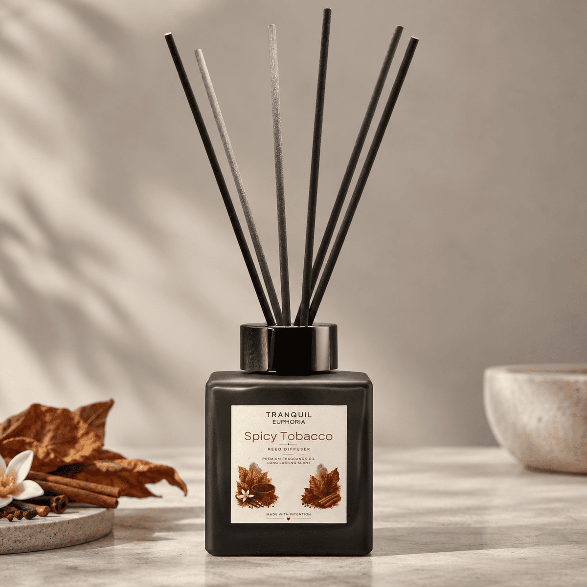 A reed diffuser bottle with six black reeds, labeled 'Tranquil Euphoria Spicy Tobacco', set on a light marble surface with dried leaves and a ceramic bowl in the background.