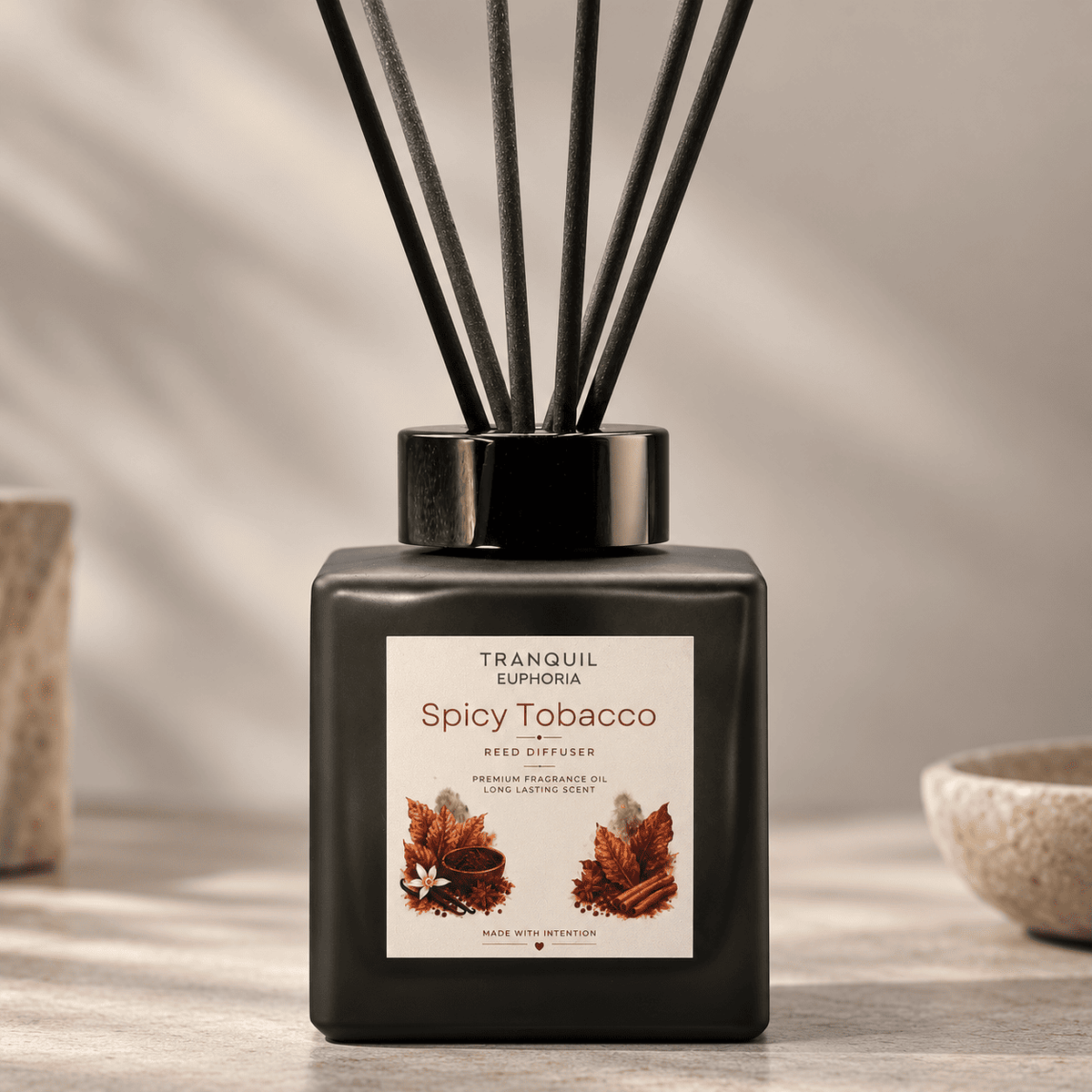 A reed diffuser bottle labeled Tranquil Euphoria Spicy Tobacco, with a matte black glass body, black cap, and dark reeds, set on a tabletop with soft shadows and decorative bowls in the background.