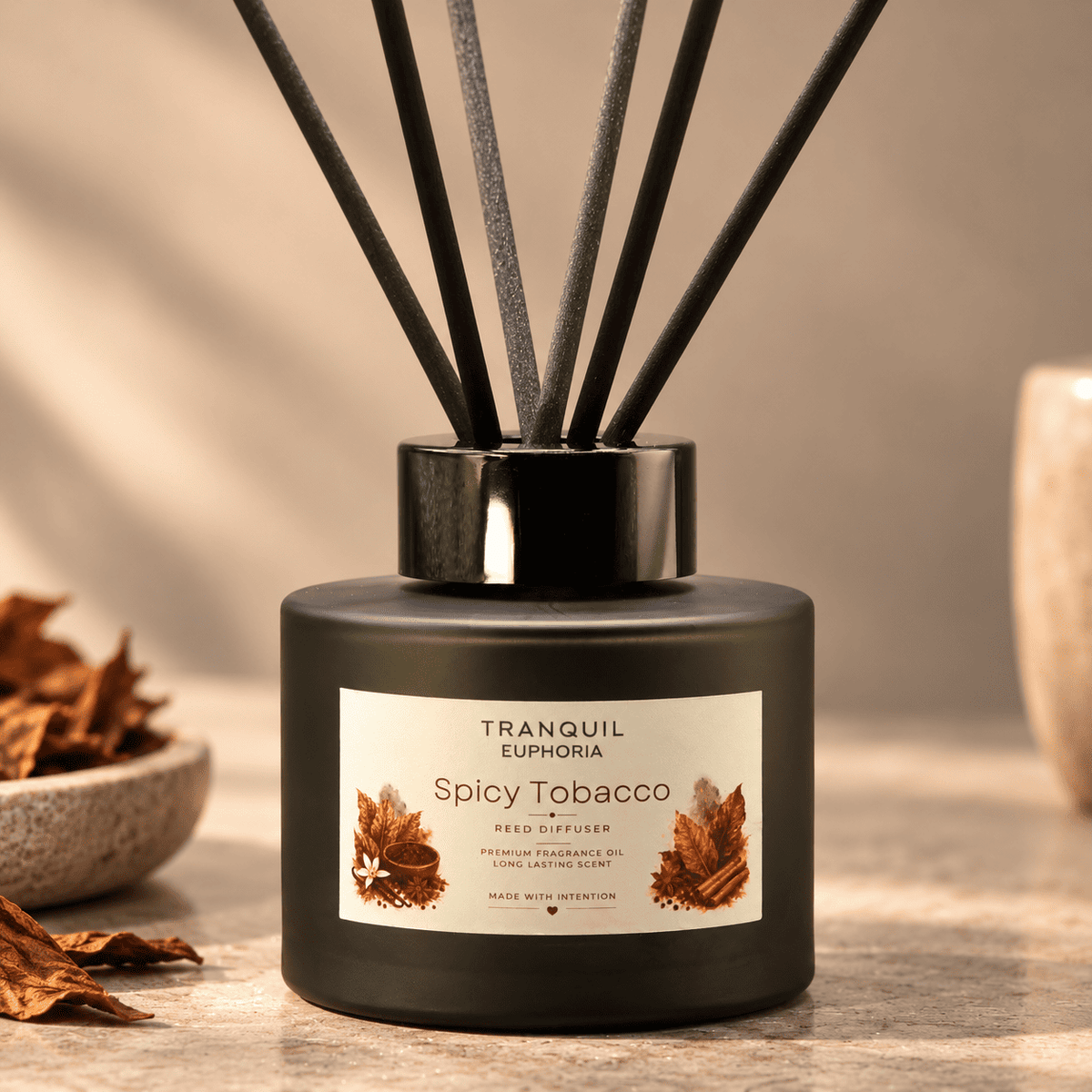 A reed diffuser bottle labeled 'Tranquil Euphoria Spicy Tobacco' with dark reeds, set on a tabletop with dried leaves and a softly lit neutral background.