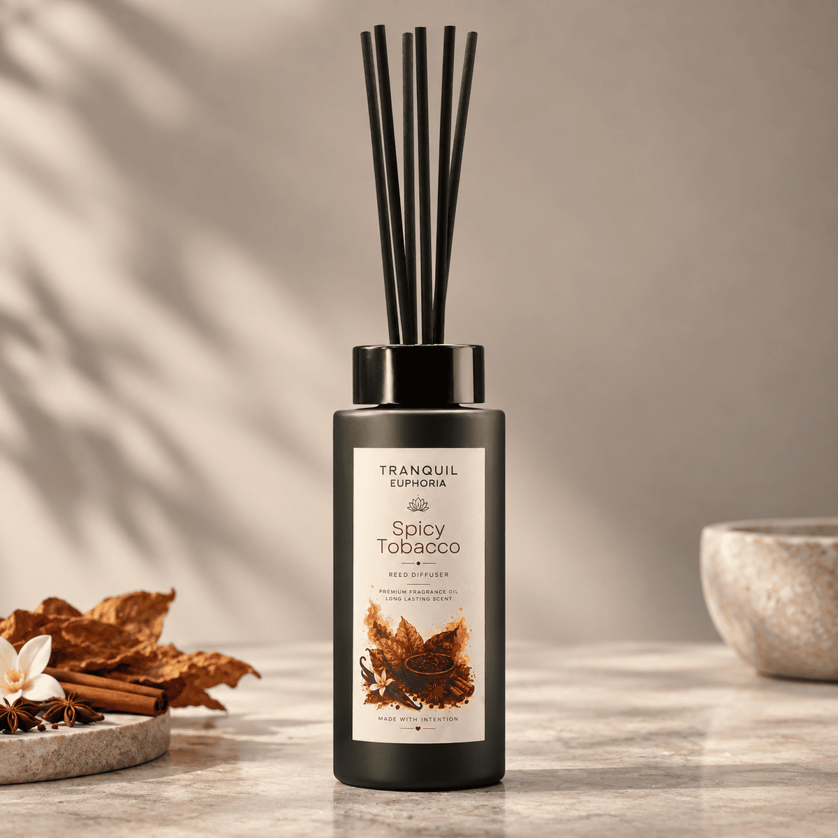 A reed diffuser bottle with black reeds stands on a stone surface in a styled interior scene. The bottle is matte black with a cream label reading Tranquil Euphoria and Spicy Tobacco, and the scene includes dried leaves, a flower, cinnamon sticks, and a small bowl in the background.