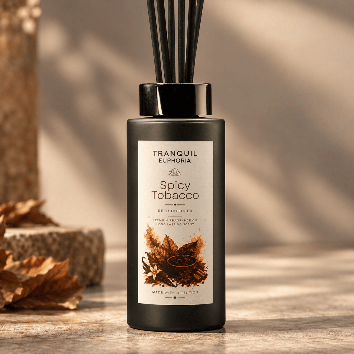 A black glass reed diffuser bottle labeled 'Tranquil Euphoria Spicy Tobacco' with dark reeds, set on a wooden surface with warm autumn leaves in the background.