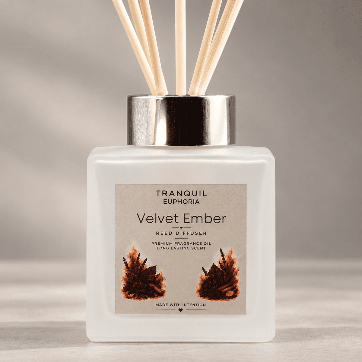 A reed diffuser bottle with a frosted white glass body, a metallic silver cap, and natural reed sticks. The front label reads 'TRANQUIL EUPHORIA Velvet Ember' with two orange-brown feather illustrations on a neutral beige background.