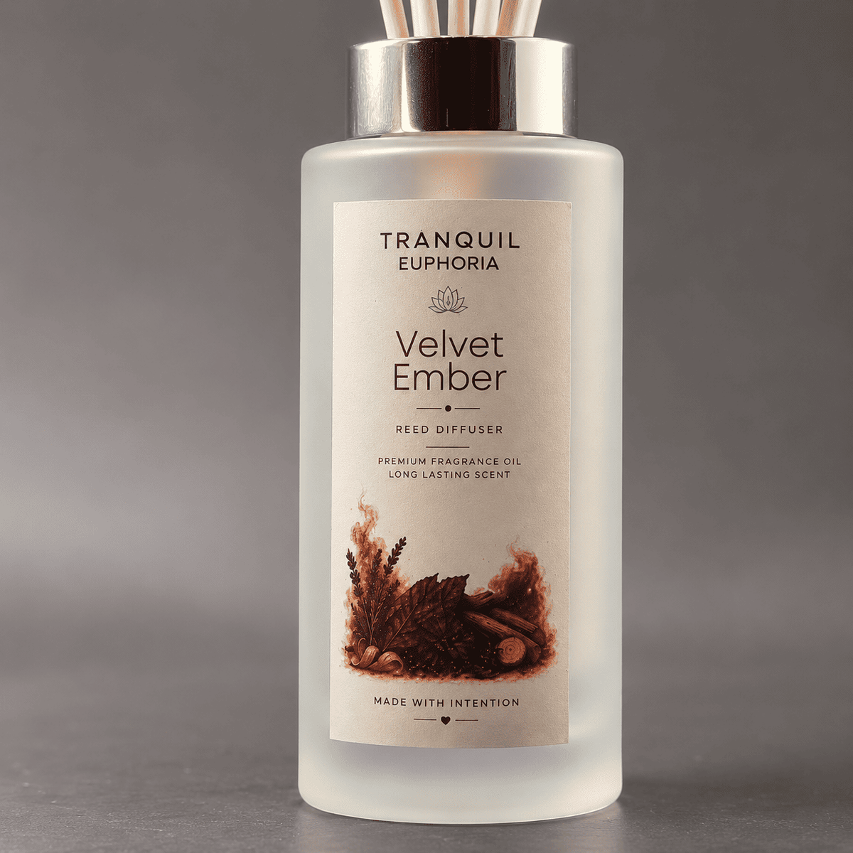 A frosted glass reed diffuser bottle with a metallic silver cap and wooden reeds, featuring a cream label with 'TRANQUIL EUPHORIA Velvet Ember' and a warm brown illustration of dried botanicals.