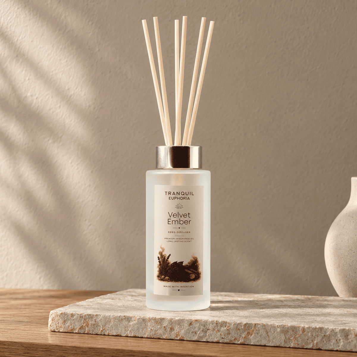 A Tranquill Euphoria Velvet Ember reed diffuser bottle with a frosted glass body, silver cap, and natural wood reeds, placed on a stone slab in a warm, softly lit room.