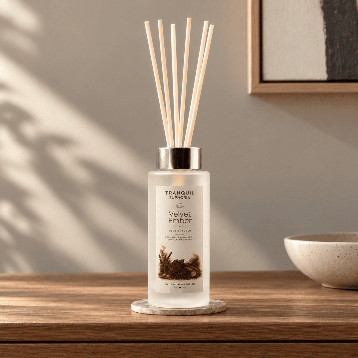 A Tranquil Euphoria Velvet Ember reed diffuser bottle with a silver cap and natural wood reeds, sitting on a marble coaster on a wooden table in a styled interior setting.