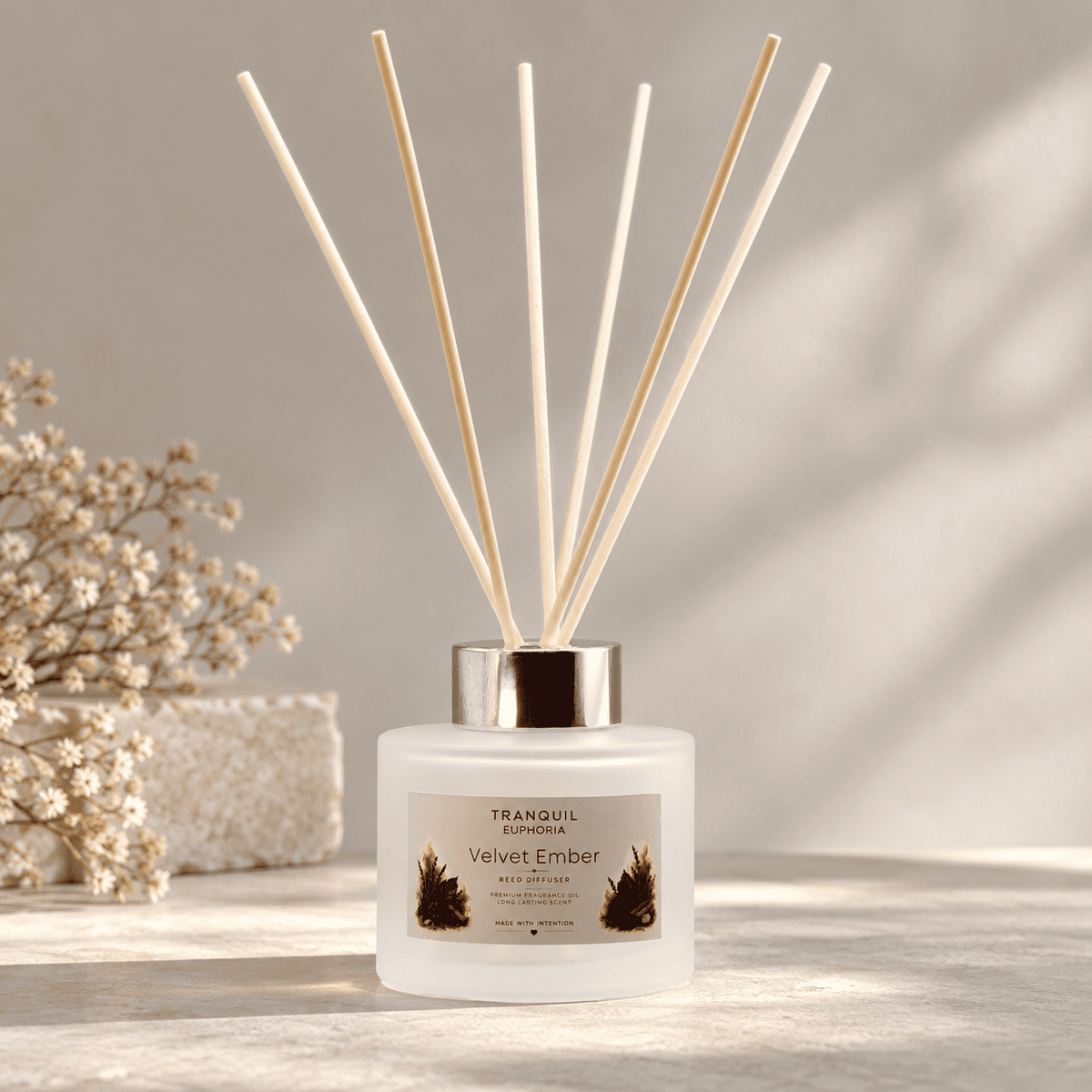 A frosted glass reed diffuser bottle with a silver cap and six natural wood reeds, labeled Tranquil Euphoria Velvet Ember, styled on a light stone surface with dried flowers in soft shadow.