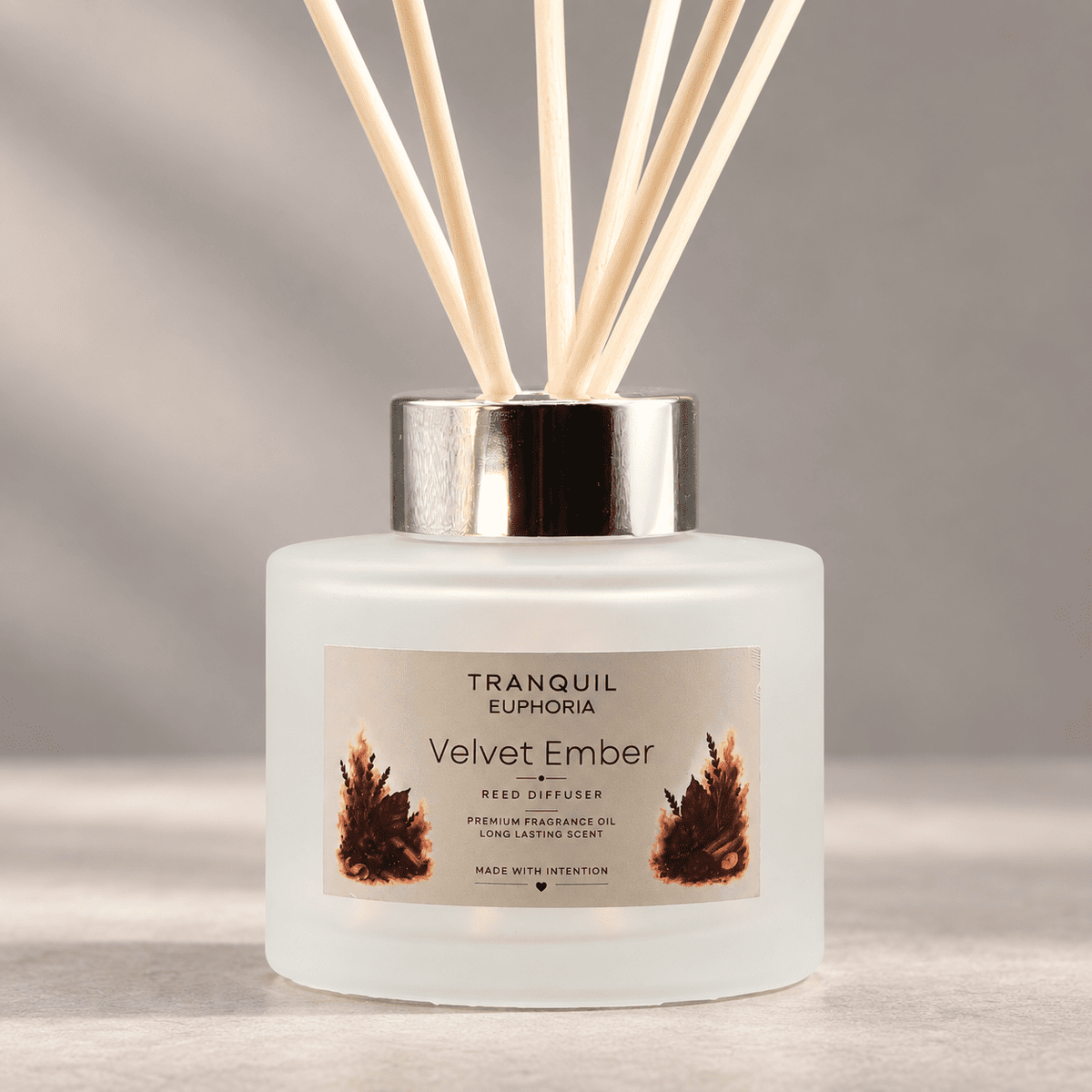 A frosted glass reed diffuser bottle with a silver cap and natural reed sticks, labeled 'Tranquil Euphoria Velvet Ember' on a soft gray background.