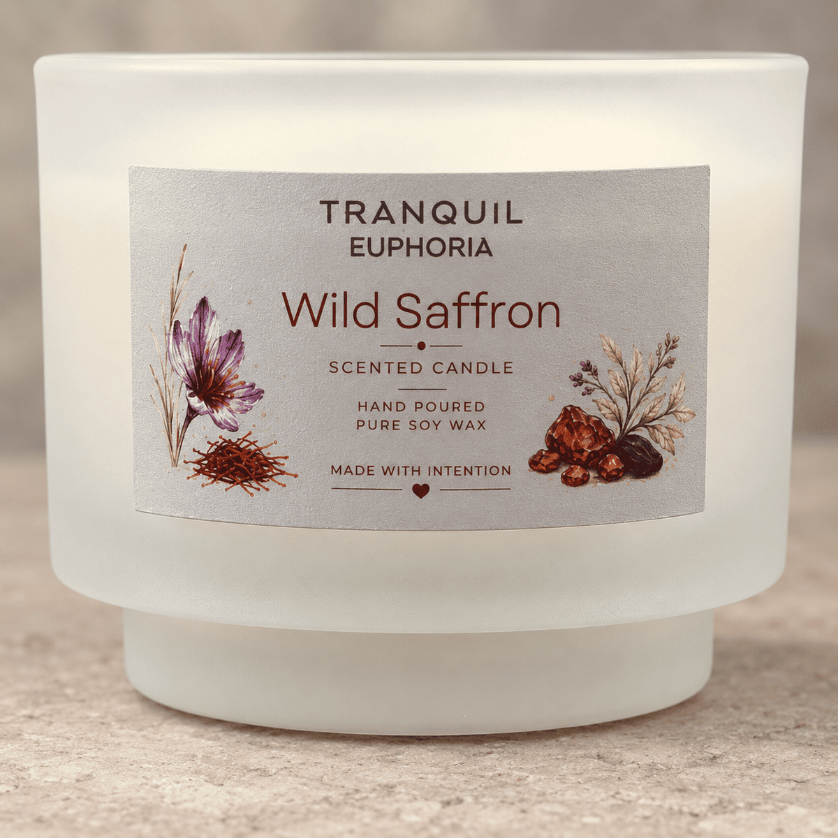 A frosted glass jar candle labeled 'Tranquil Euphoria Wild Saffron' with botanical illustrations on the front, photographed on a neutral stone-textured surface.