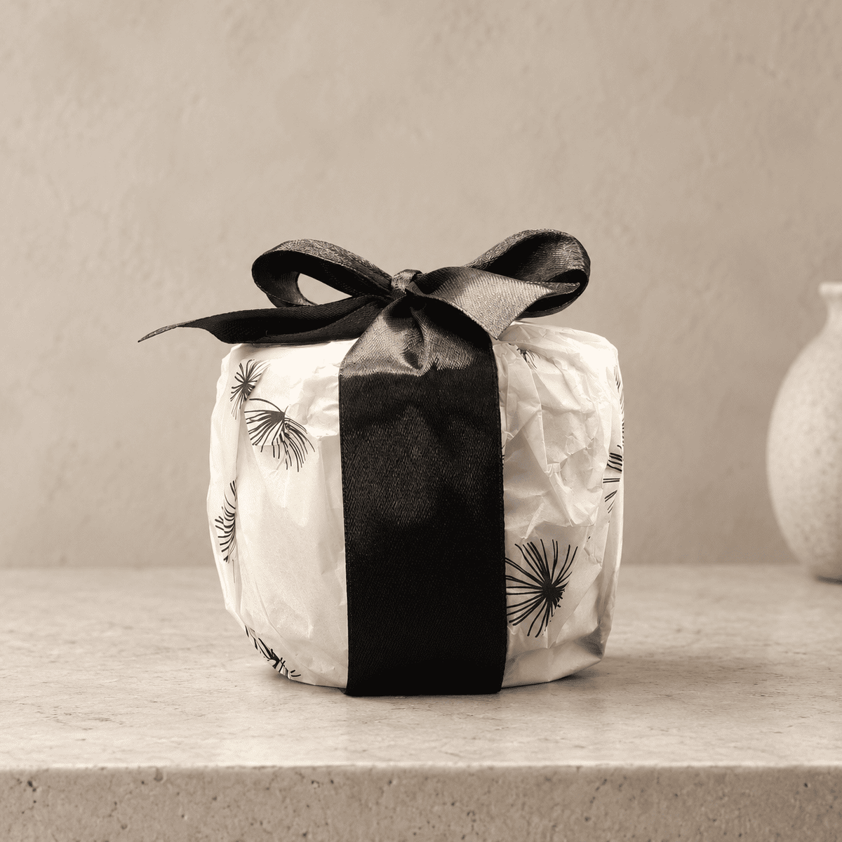 A small wrapped Wild Saffron candle package wrapped in off-white paper with a black dandelion print and tied with a wide black satin ribbon bow, set on a light stone surface against a warm beige wall.